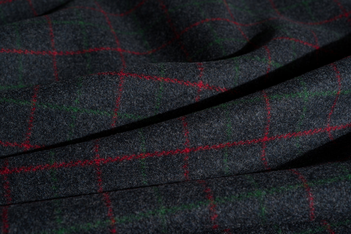 A close-up of dark charcoal gray wool fabric with a subtle woven texture, overlaid with a red and green windowpane plaid pattern.