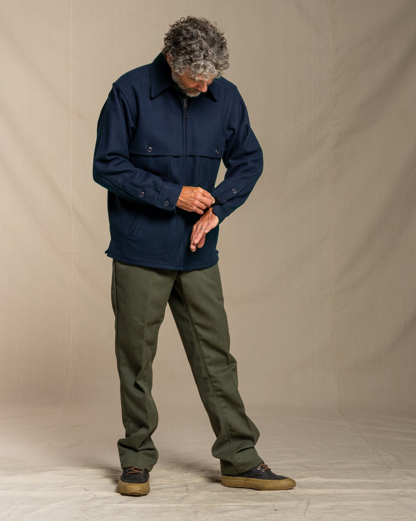 Man wearing a navy jacket and green pants against a beige background