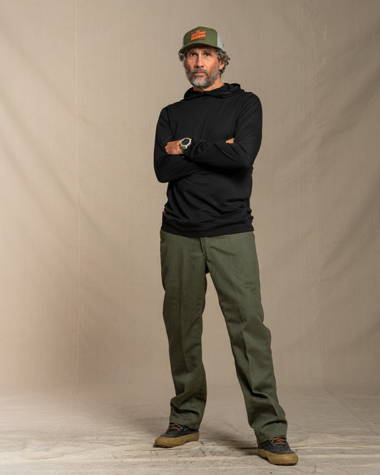 Man wearing a black merino wool hoodie, green cap, and green pants standing against a beige background