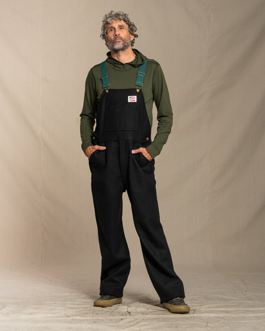 Man wearing wool black bib overalls and green hoodie against a plain background