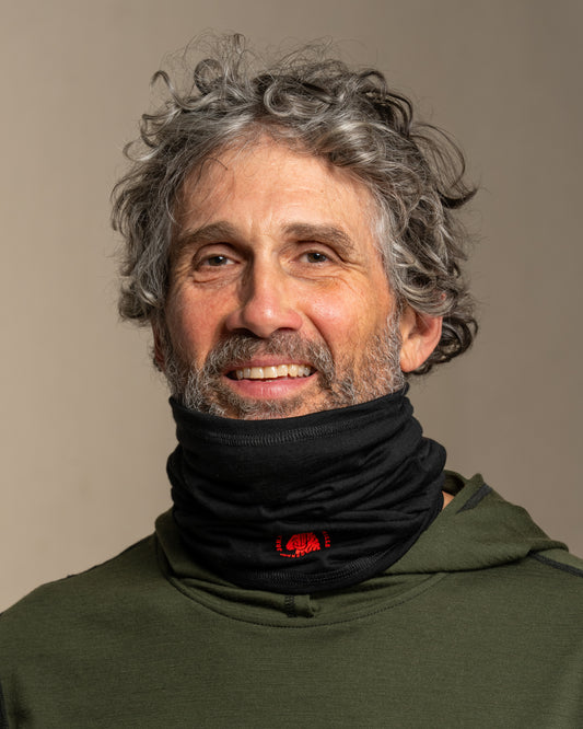Man wearing a green hoodie and black merino wool neck gaiter with a red logo against a beige background