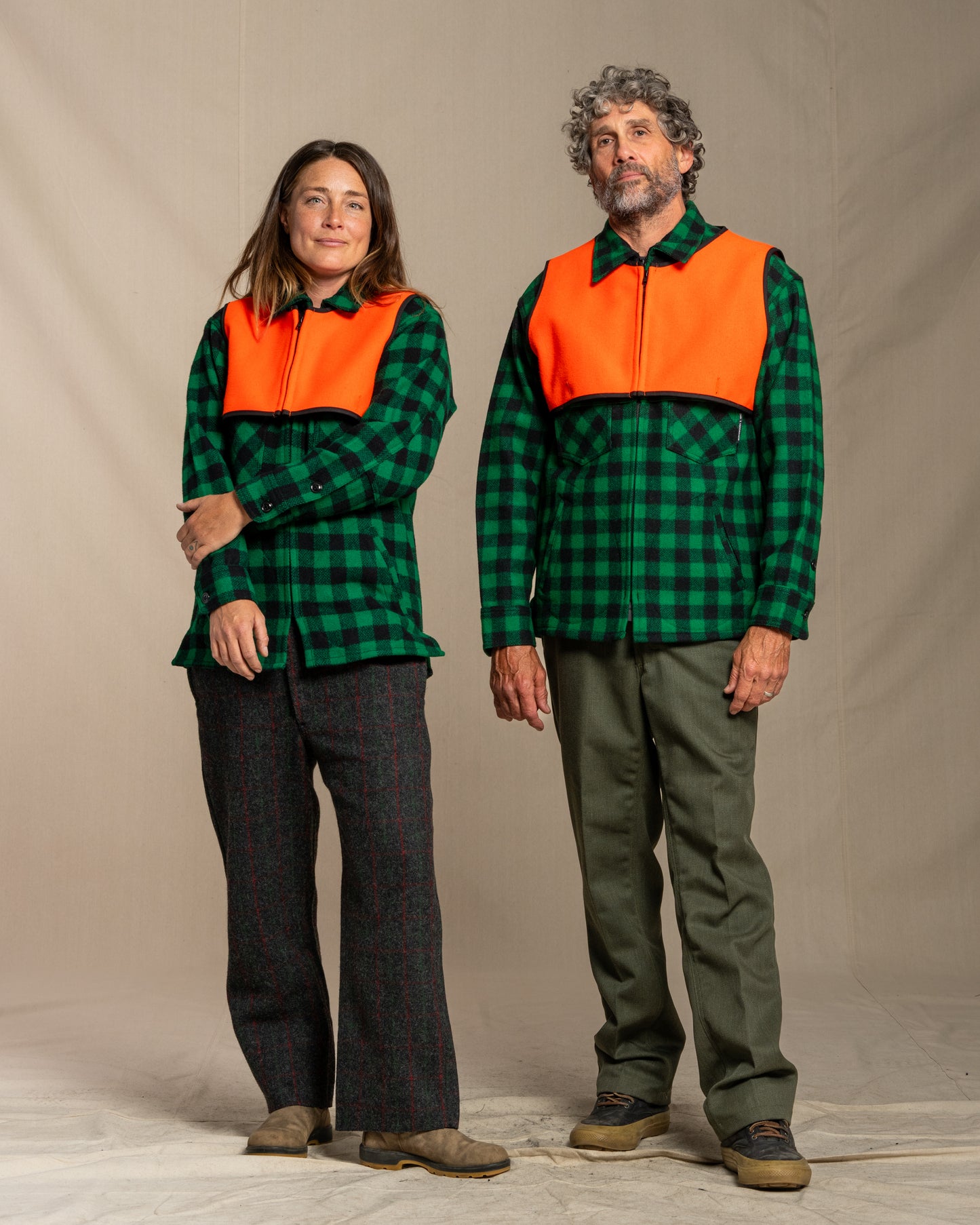 Two people wearing green checkered jackets with blaze orange wool safety vests against a neutral background