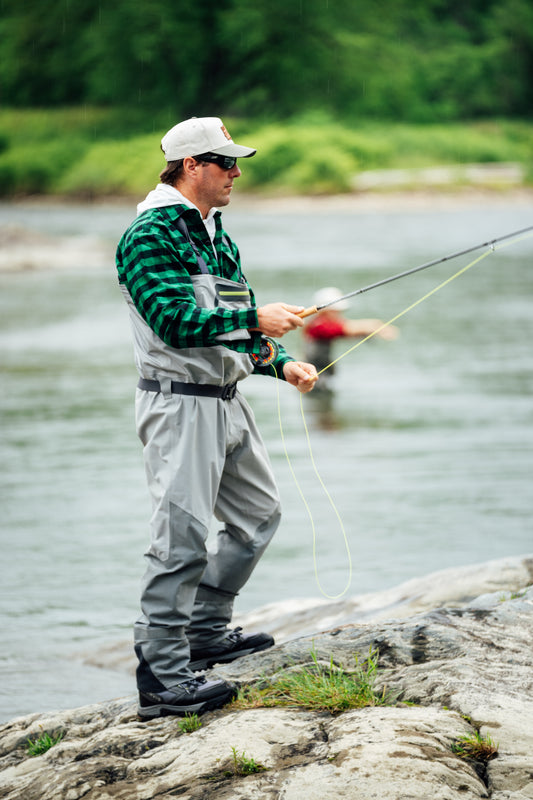 A man fly fishing in a river wearing gray waders, a green and black plaid shirt, and a white cap, standing on a rocky bank with water and green trees in the background.