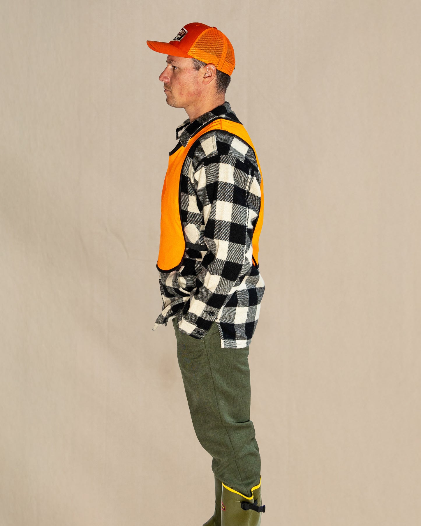 Person wearing a plaid shirt, orange vest, and green pants against a beige background