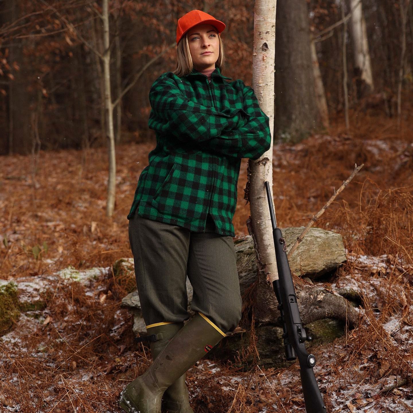woman hunter resting on a birch tree wearing wool pants, green northwoods jac shirt, orange darn tough hat