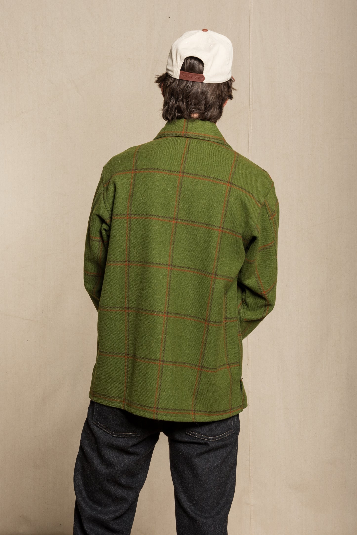A person shown from the back wearing an olive green wool-blend plaid jacket with thin red windowpane lines, paired with dark jeans and a white baseball cap with a brown leather band.