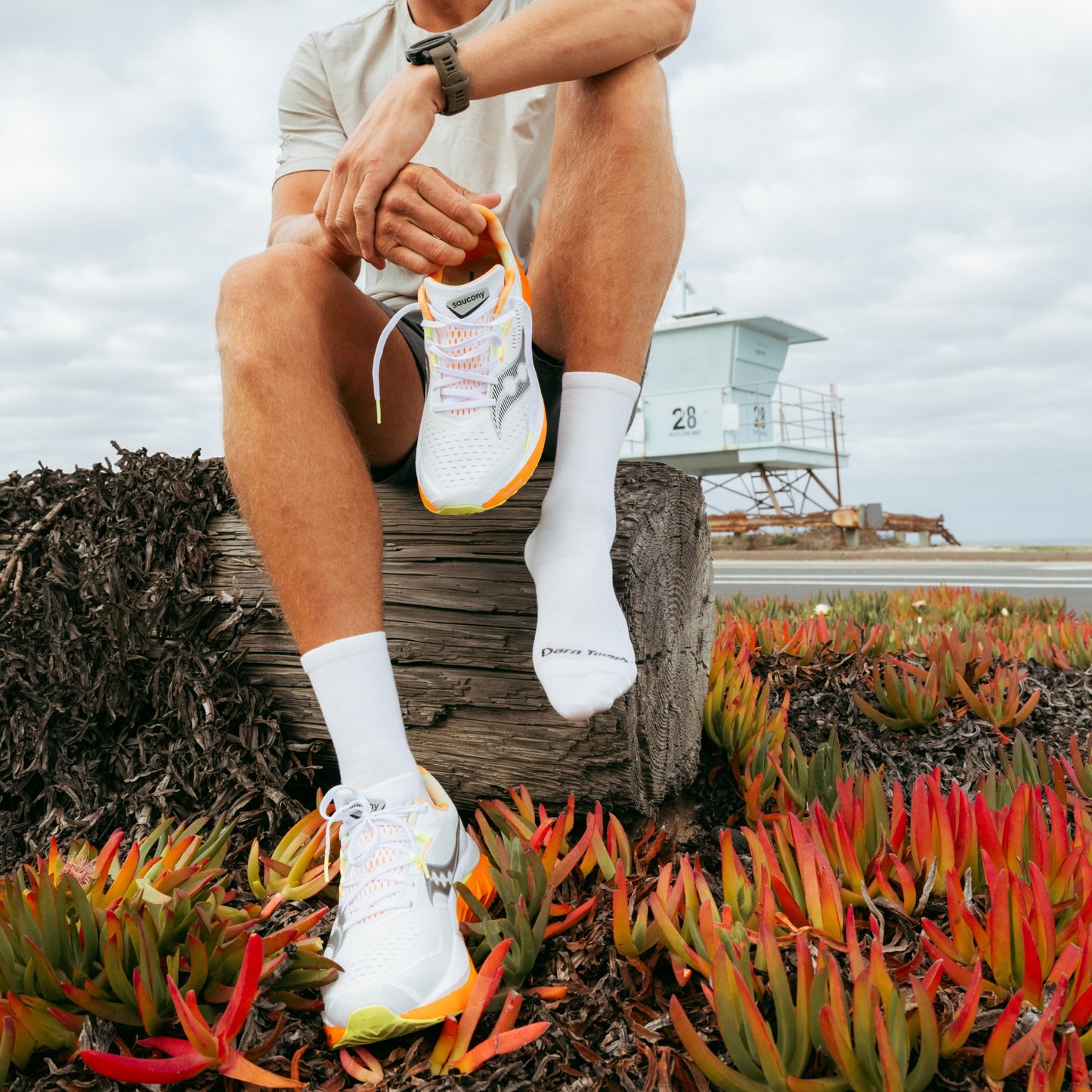 Person sitting on a log by a coastal area wearing white sneakers with orange accents.