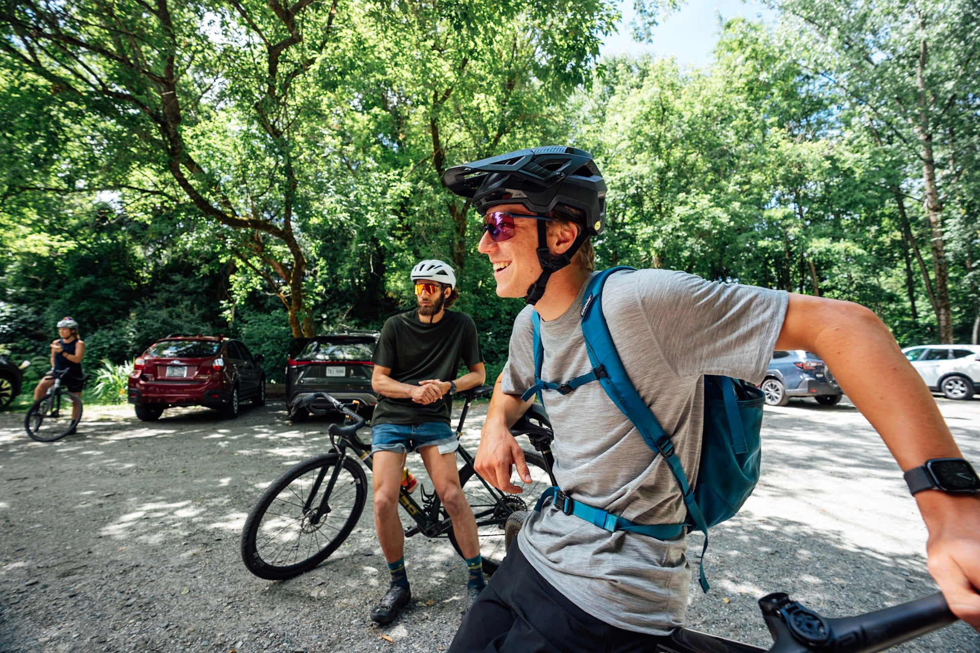 A man in a black bicycle helmet and sunglasses rides a mountain bike in a wooded outdoor area, wearing a gray merino wool t-shirt and a blue backpack. Another cyclist stands on a bike nearby, with trees, parked cars, and other riders visible in the background.