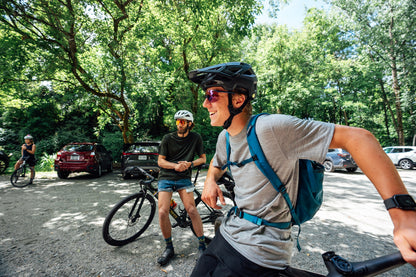 A man in a black bicycle helmet and sunglasses rides a mountain bike in a wooded outdoor area, wearing a gray merino wool t-shirt and a blue backpack. Another cyclist stands on a bike nearby, with trees, parked cars, and other riders visible in the background.