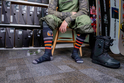 Darn Tough Men's Thermolite Beer Badge Over-the-Calf Midweight Ski & Snowboard Sock on model sitting on chair