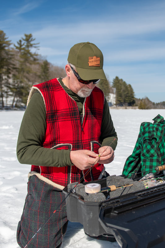 A man kneeling in a snowy outdoor setting, wearing a red and black plaid vest over an olive green shirt, an olive green cap with a logo patch, and sunglasses. He is handling fishing line near an open black tackle case, with a green plaid shirt draped over the side of the case.