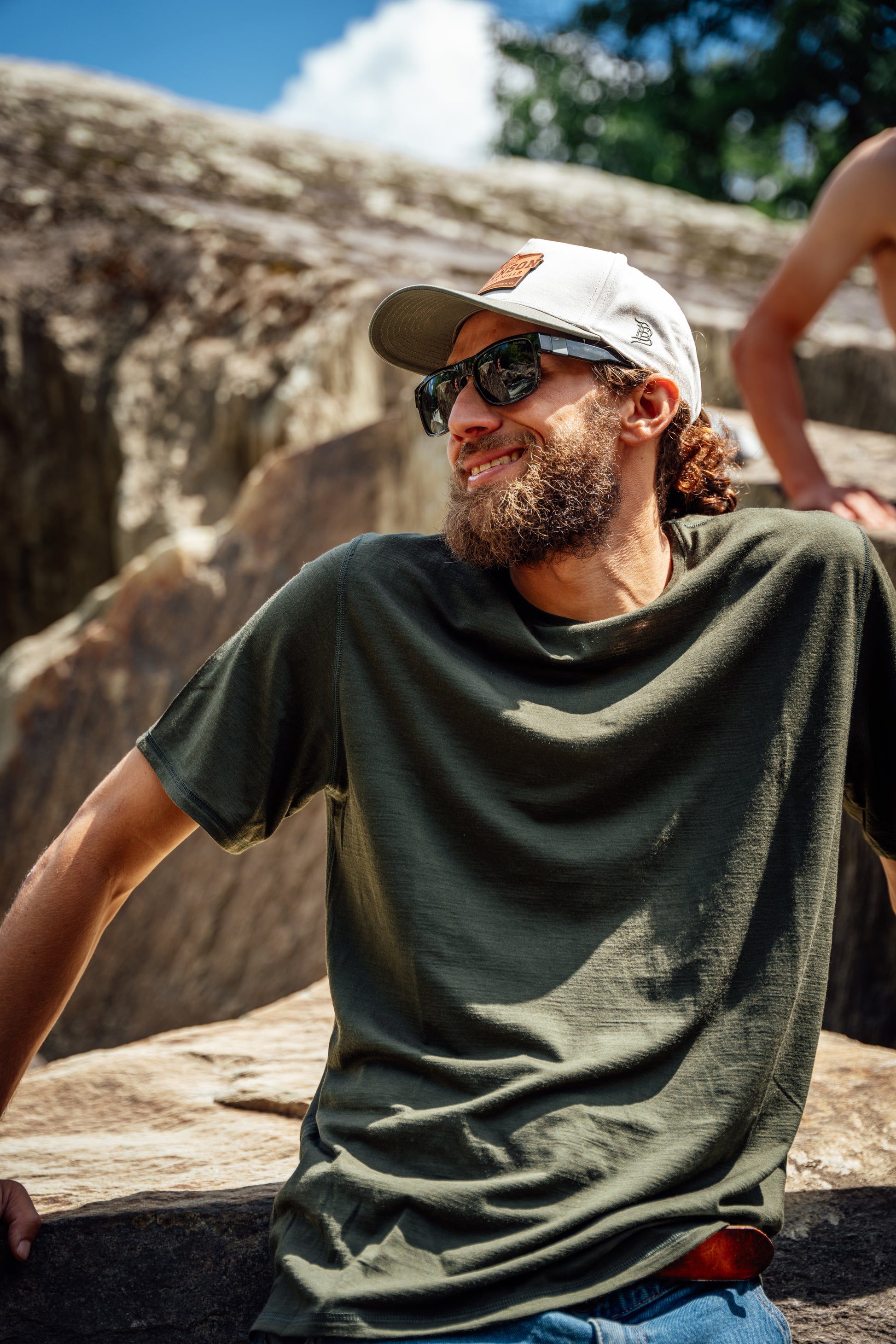 A man wearing an olive green merino wool t-shirt, a white baseball cap, and sunglasses, sitting outdoors against a rocky cliff background.