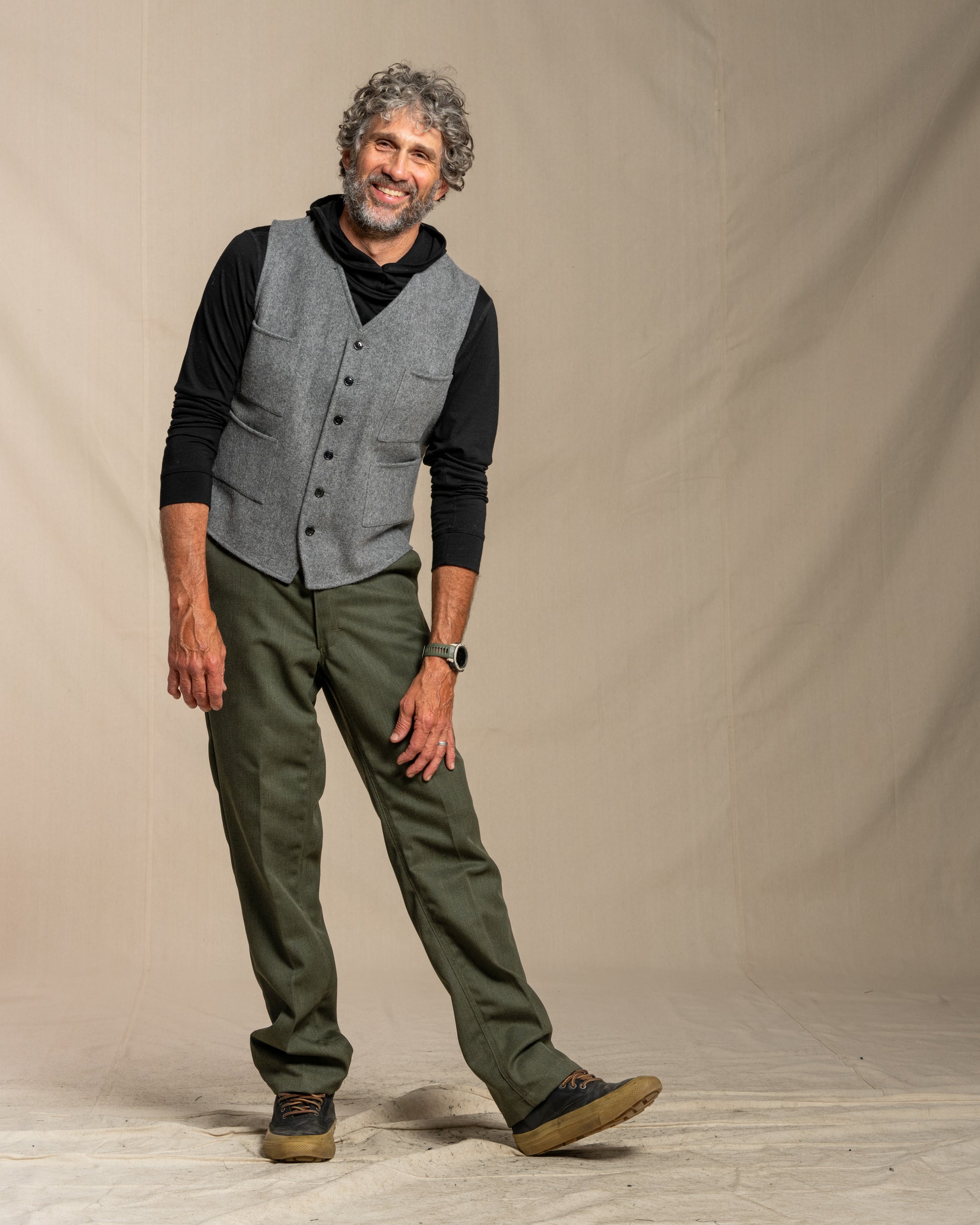 Man wearing a gray vest, black shirt, green pants, and brown boots against a beige background