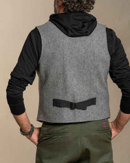 Person wearing a gray vest with black sleeves, standing against a beige background.