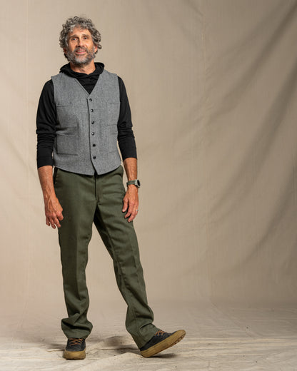 Man wearing a gray vest over a black shirt with green pants against a plain background