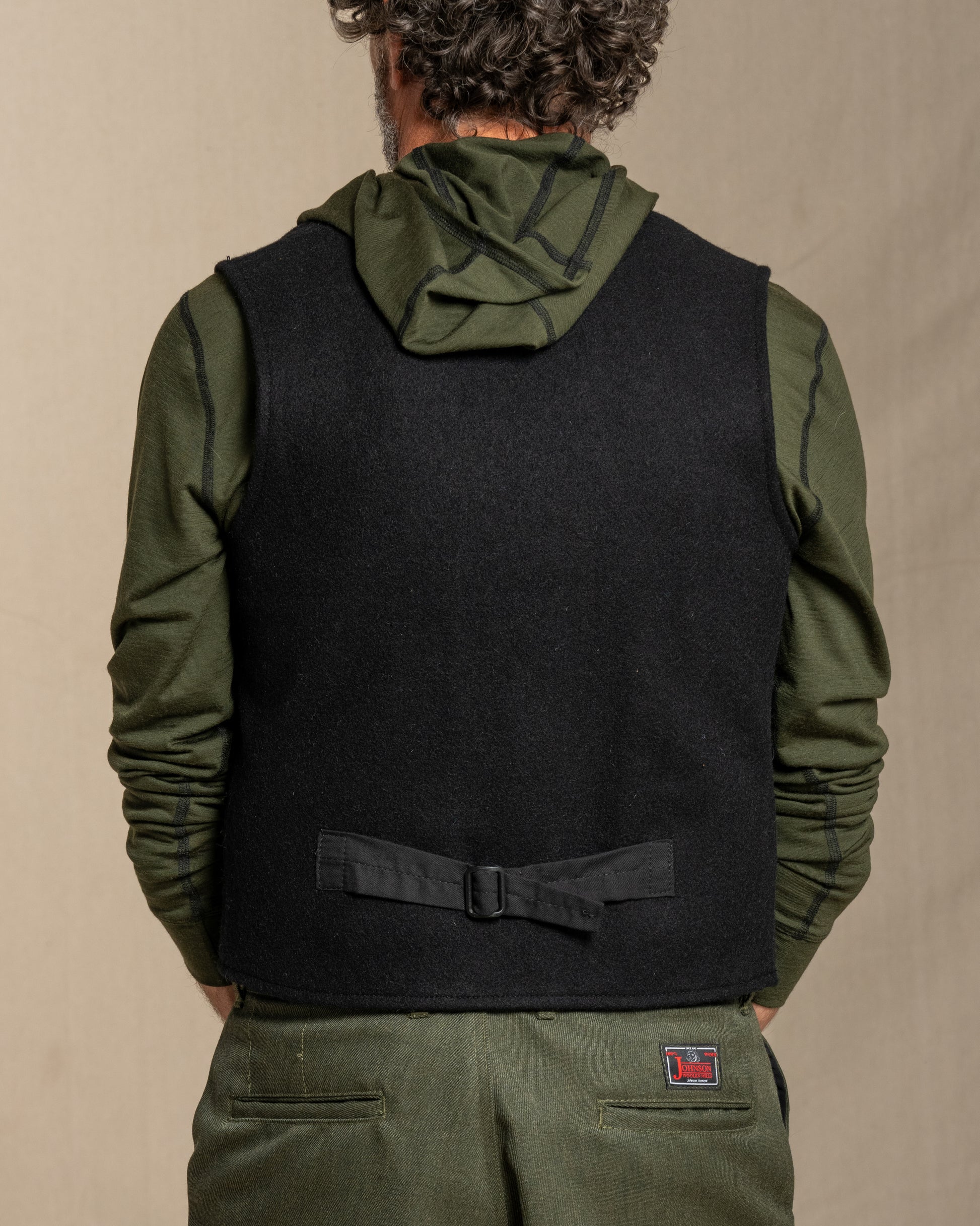 Person wearing a black vest over a green jacket with a beige background