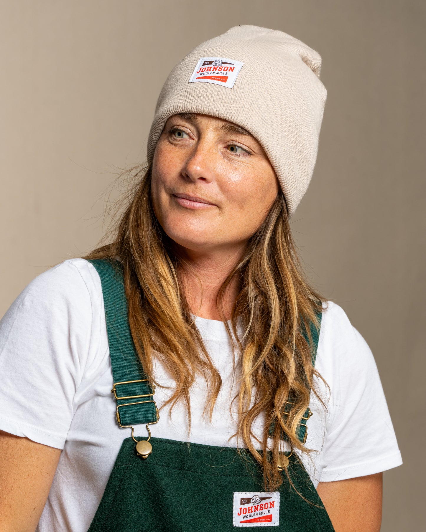 Person wearing an oatmeal beanie and green overalls against a plain background