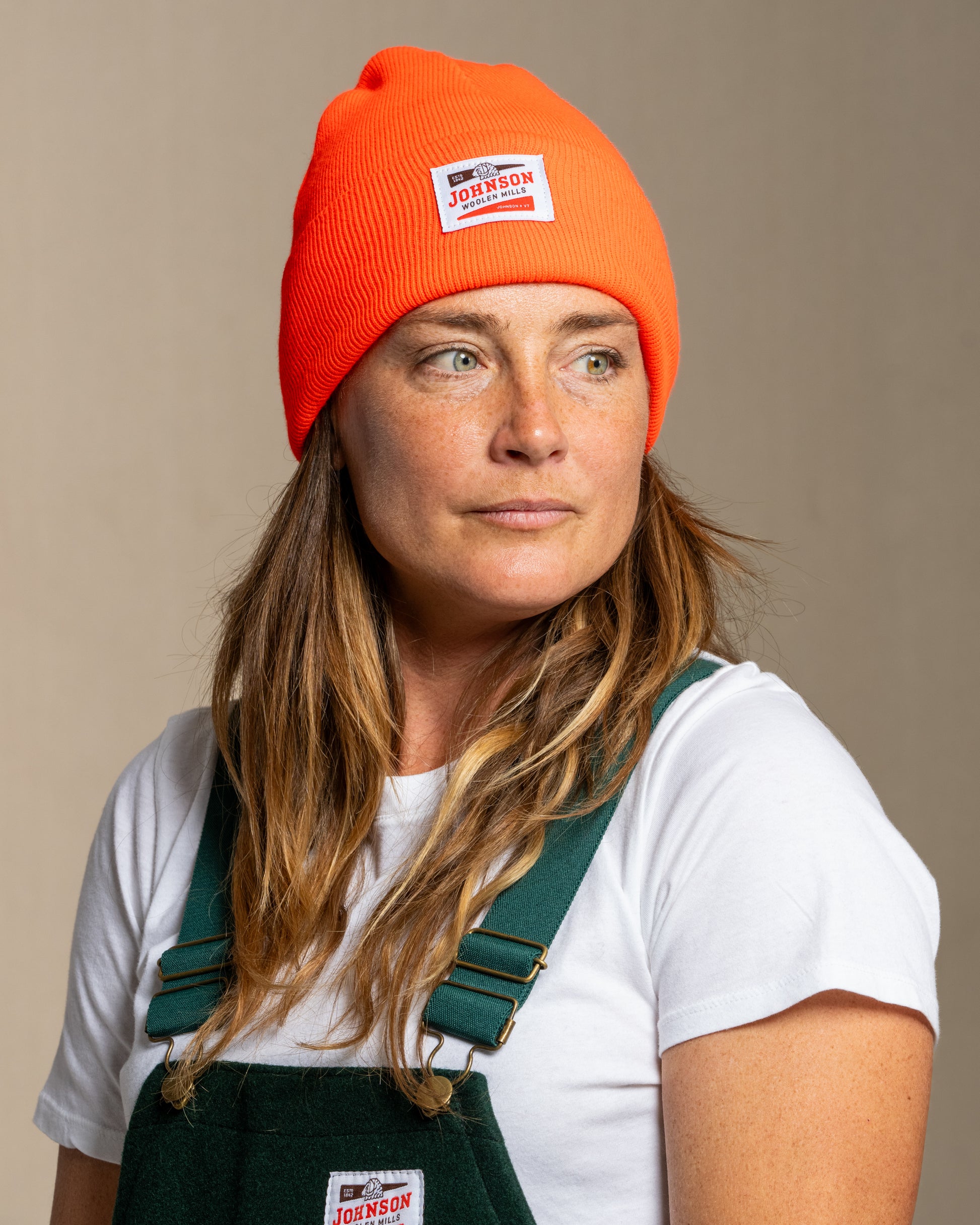 Person wearing a blaze orange beanie and green overalls with a neutral background