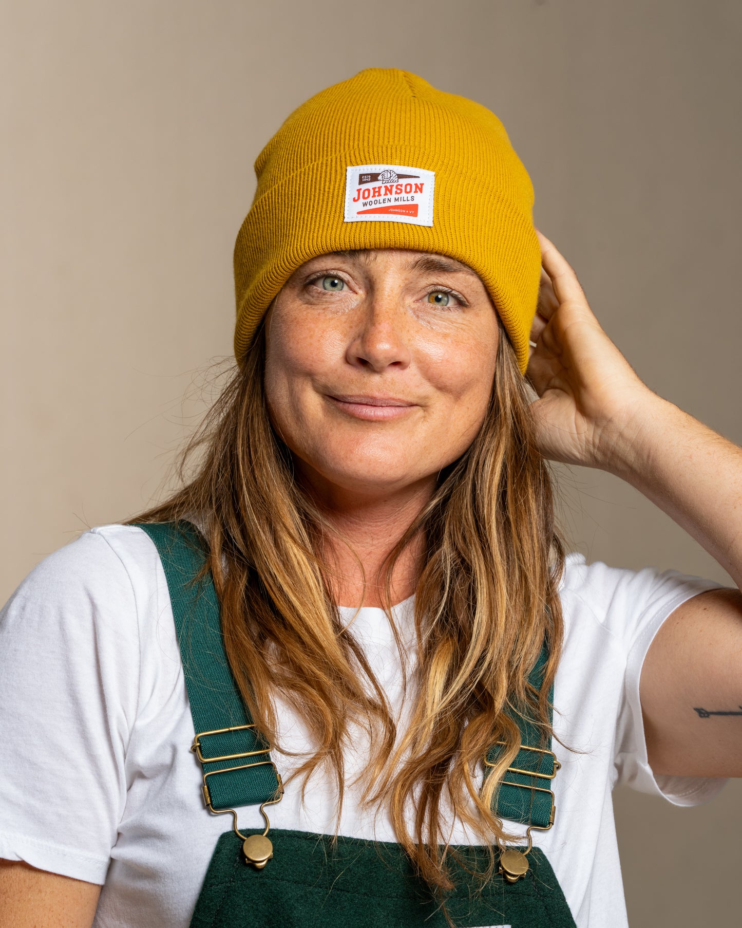 Person wearing a mustard yellow beanie with Johnson logo and green overalls against a beige background