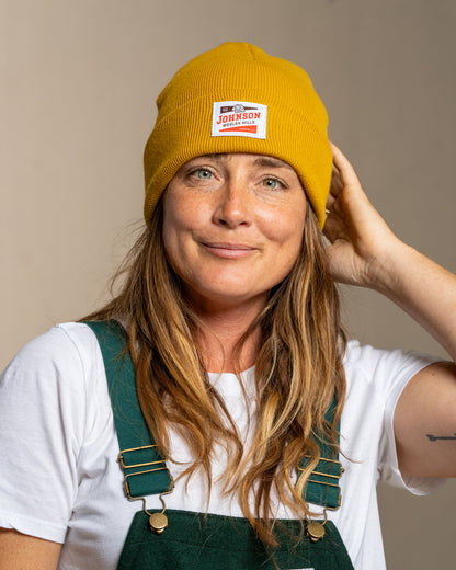 Person wearing a mustard yellow beanie with Johnson logo and green overalls against a beige background