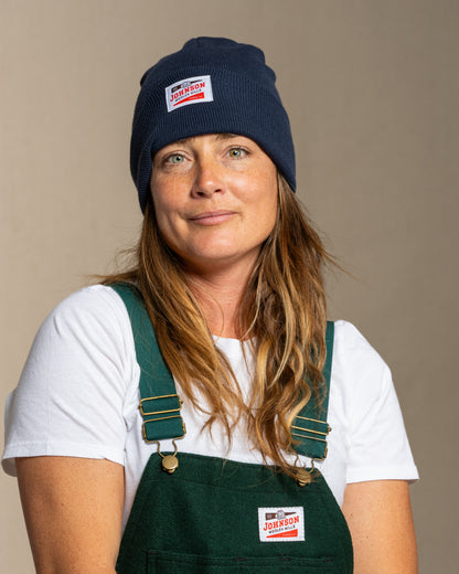 Person wearing a navy beanie and green overalls with Johnson logo against a beige background