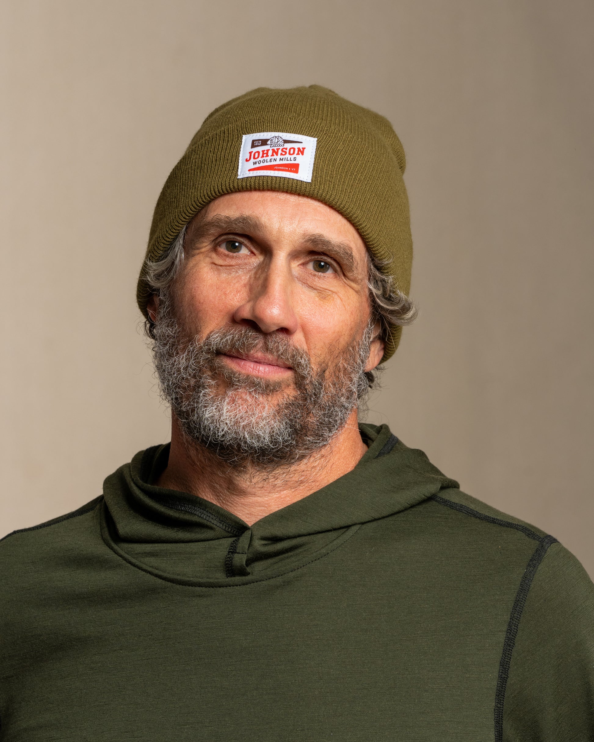 Man wearing a green beanie with Johnson logo and matching hoodie against a beige background