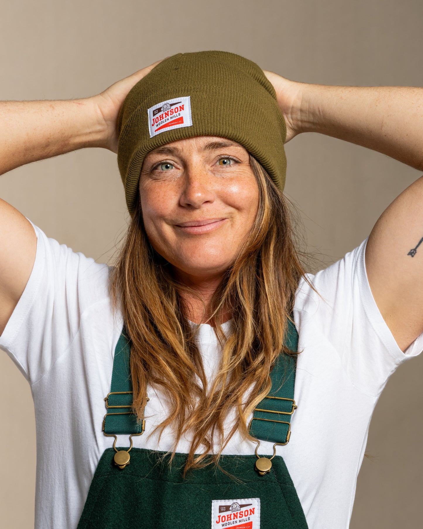 Person wearing an olive green beanie and overalls with Johnson logo against a neutral background