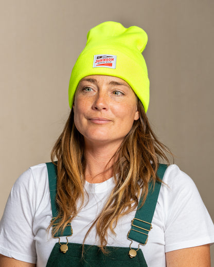 Person wearing a bright safety yellow beanie with Johnson logo and green overalls on a beige background