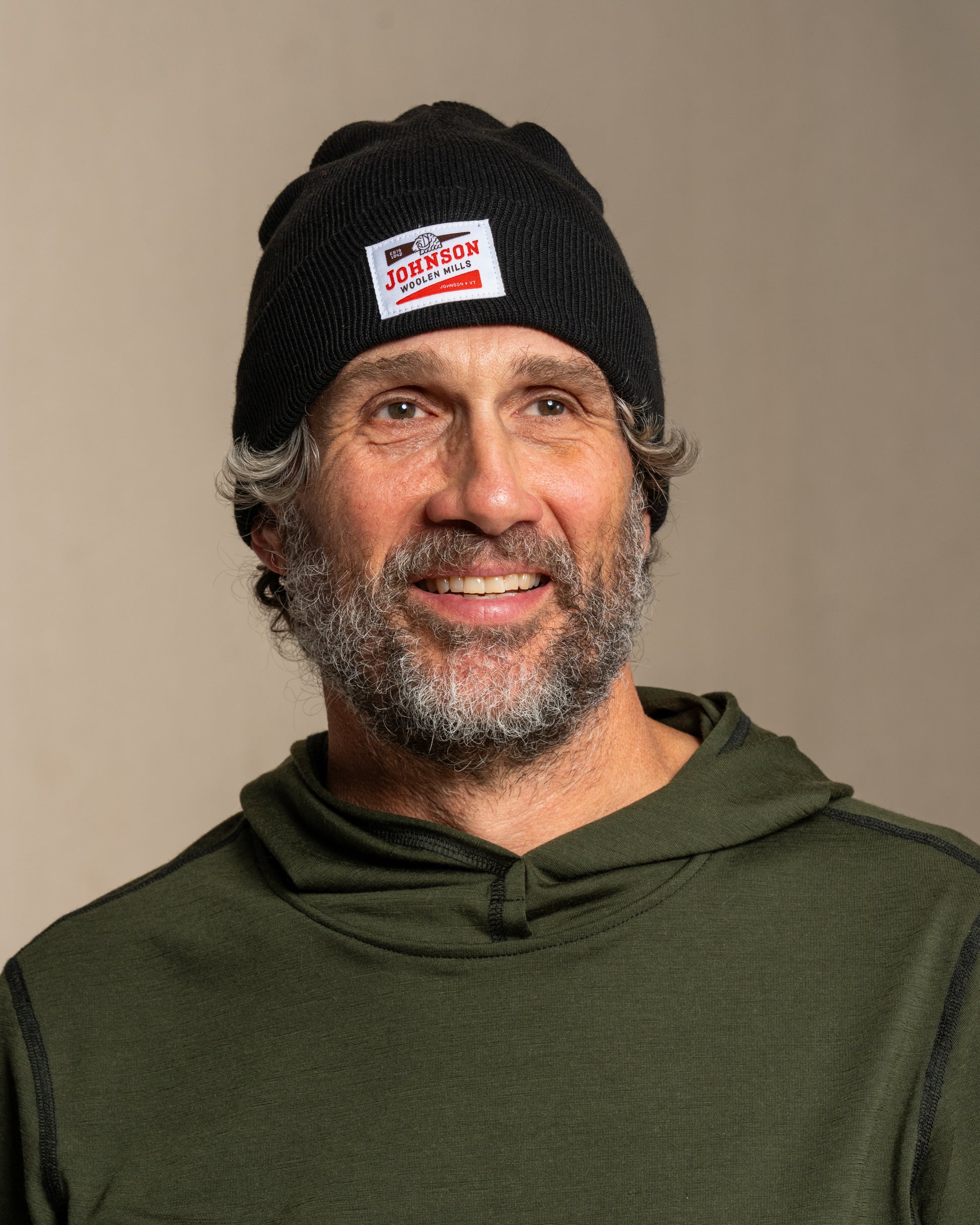 Man wearing a black beanie with Johnson logo and a green hoodie against a beige background