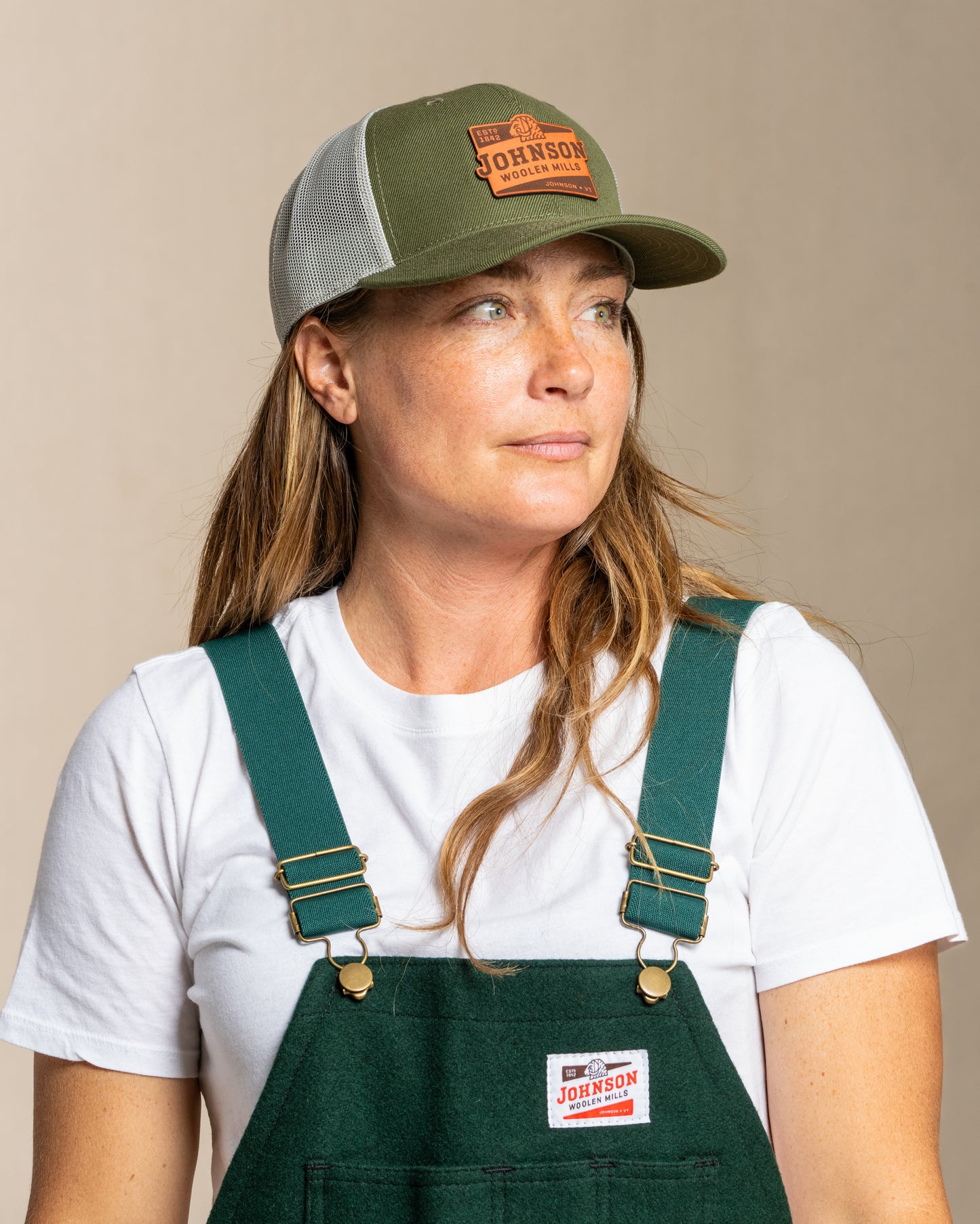 Woman wearing a green cap and green overalls with a white shirt underneath on a beige background