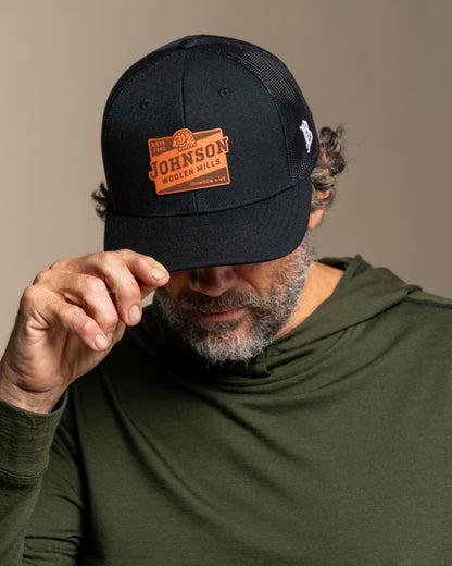 Man wearing a black cap with a Johnson logo, looking down, on a canvas background