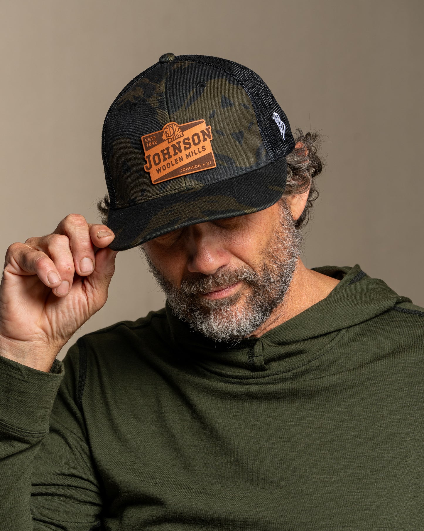 Man wearing a camouflage cap with a Johnson leather patch, posing against a canvas background