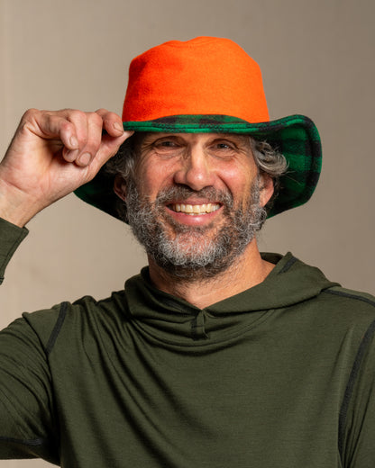 Person wearing a beagle blaze orange crusher hat with green and black brim and green merino wool hoodie against a canvas background