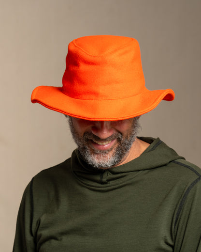 Person wearing a beagle blaze orange crusher hat and green merino wool hoodie against a canvas background