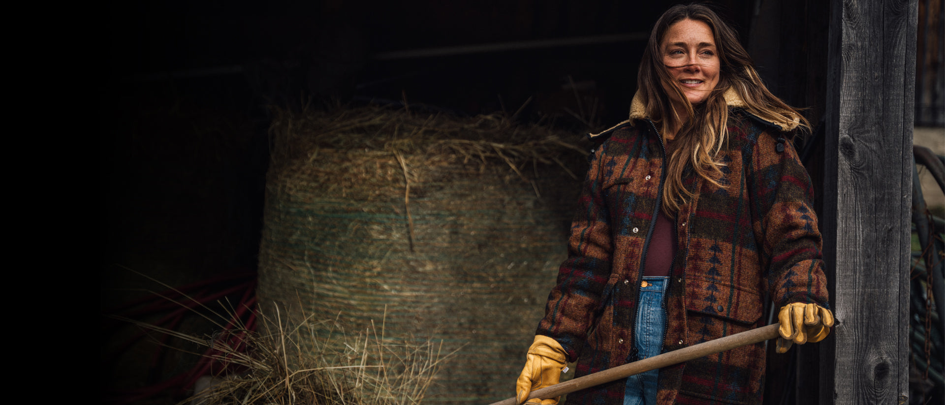 Woman in a plaid coat standing next to a hay bale in a barn.