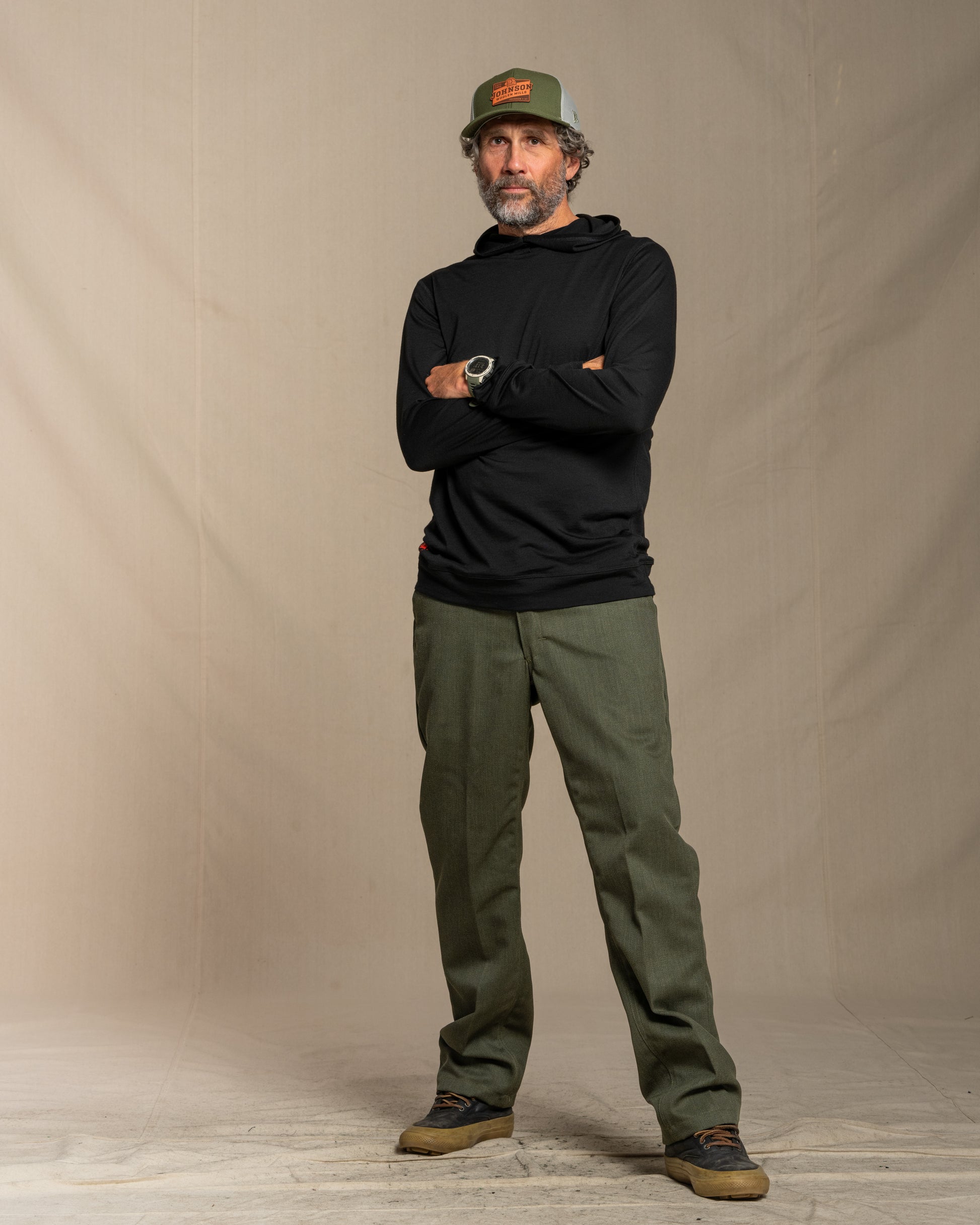 Man wearing a black merino wool hoodie, green cap, and green pants standing against a beige background
