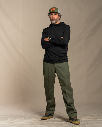Man wearing a black merino wool hoodie, green cap, and green pants standing against a beige background
