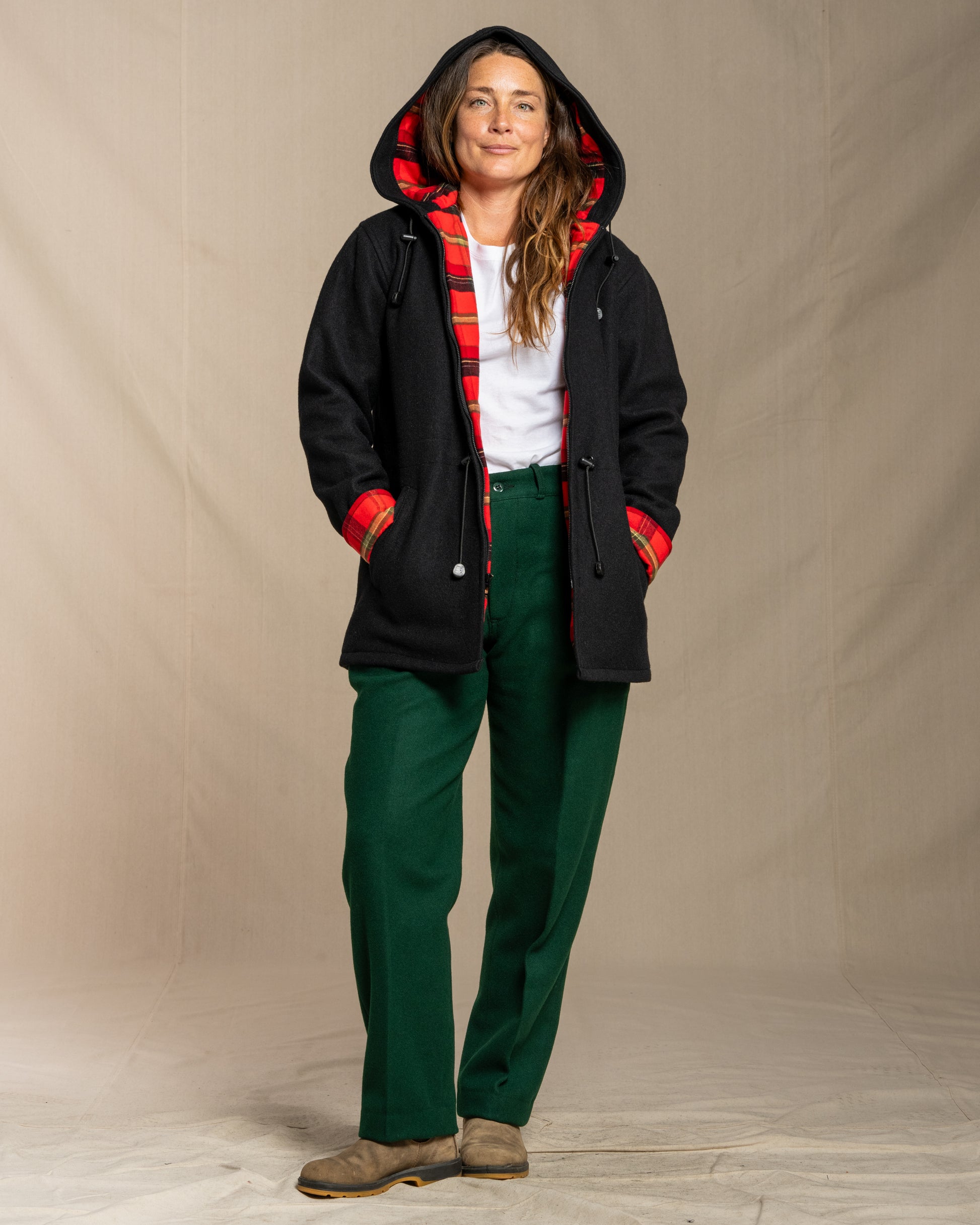 Person wearing a black wool coat with red flannel lining, white shirt, and green wool pants against a beige background