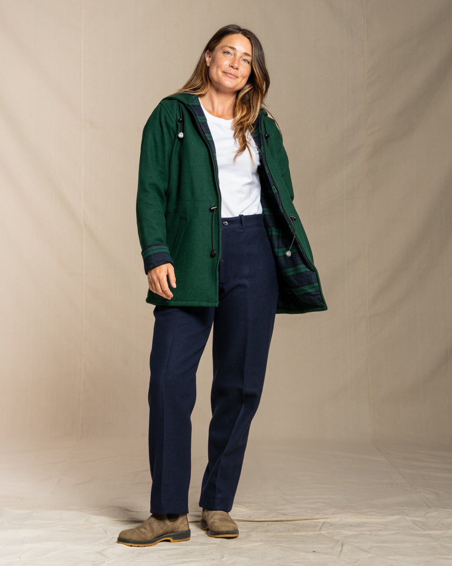 Woman wearing a green wool coat, white shirt, navy wool pants, and brown shoes against a beige background