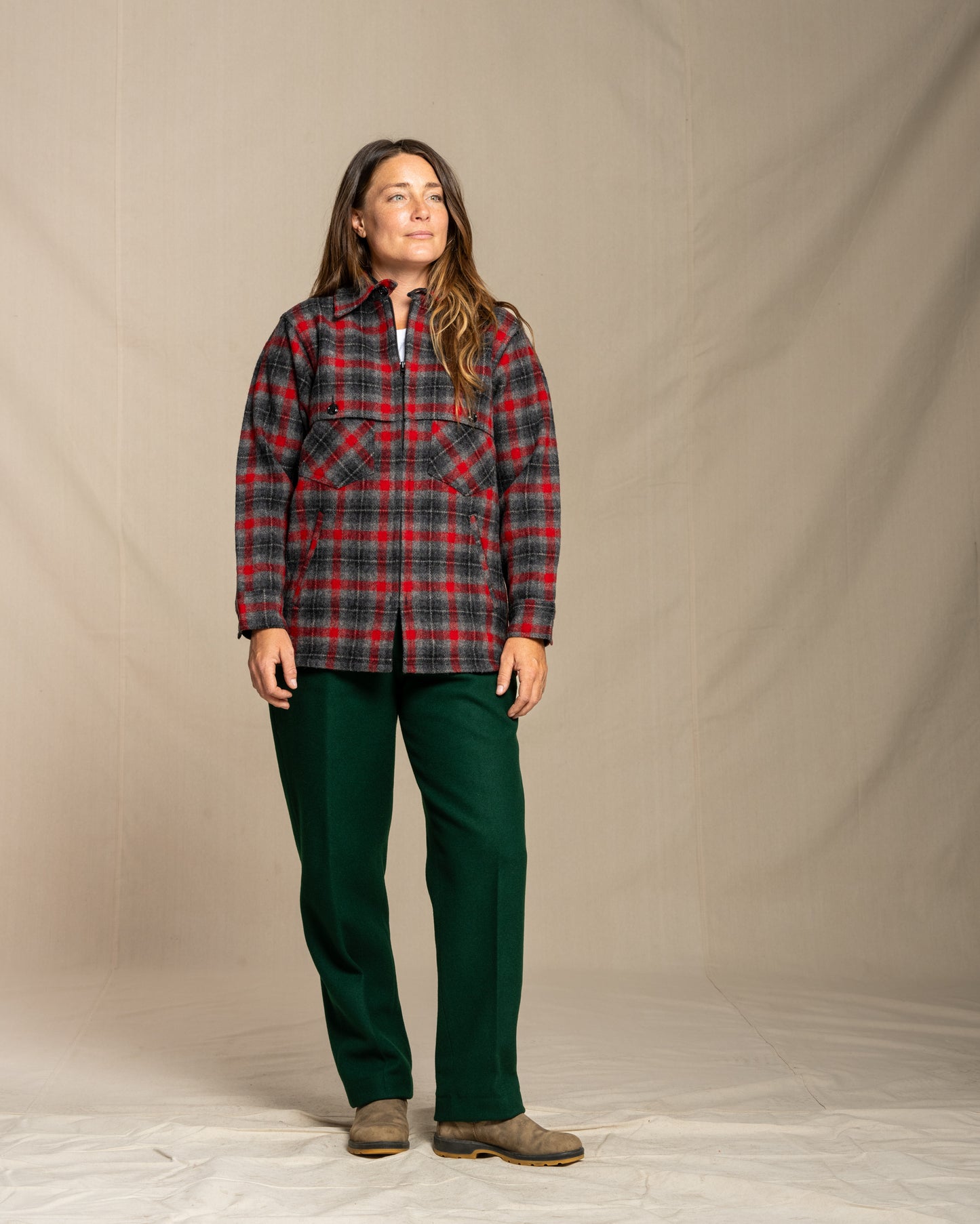 Person wearing a wool red and gray plaid jacket and green wool pants against a beige background