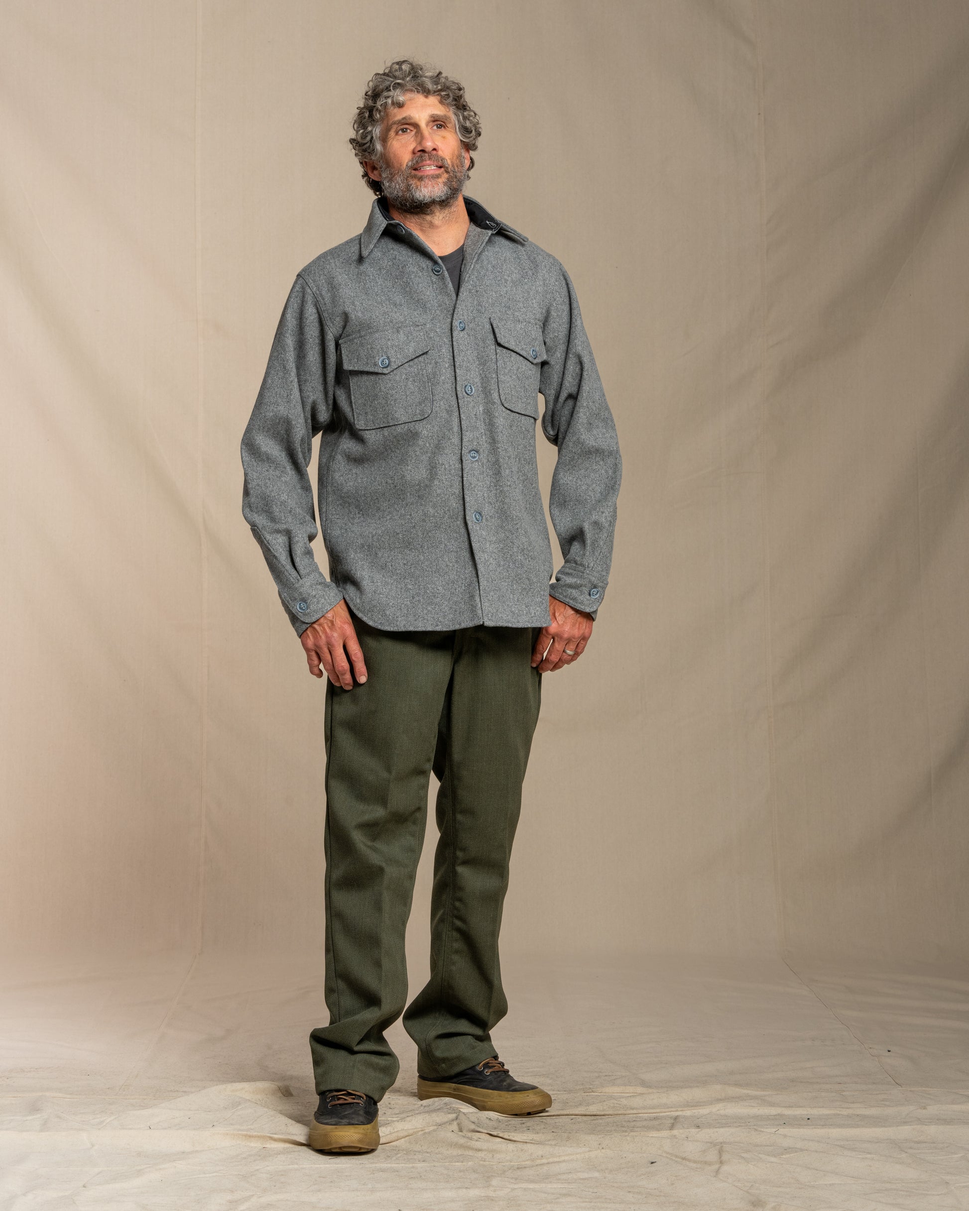 Man wearing a gray wool shirt and green pants standing against a beige background