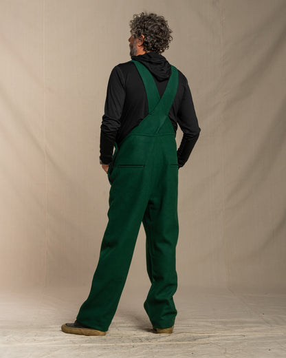 Person wearing green wool overalls and a black jacket against a beige background