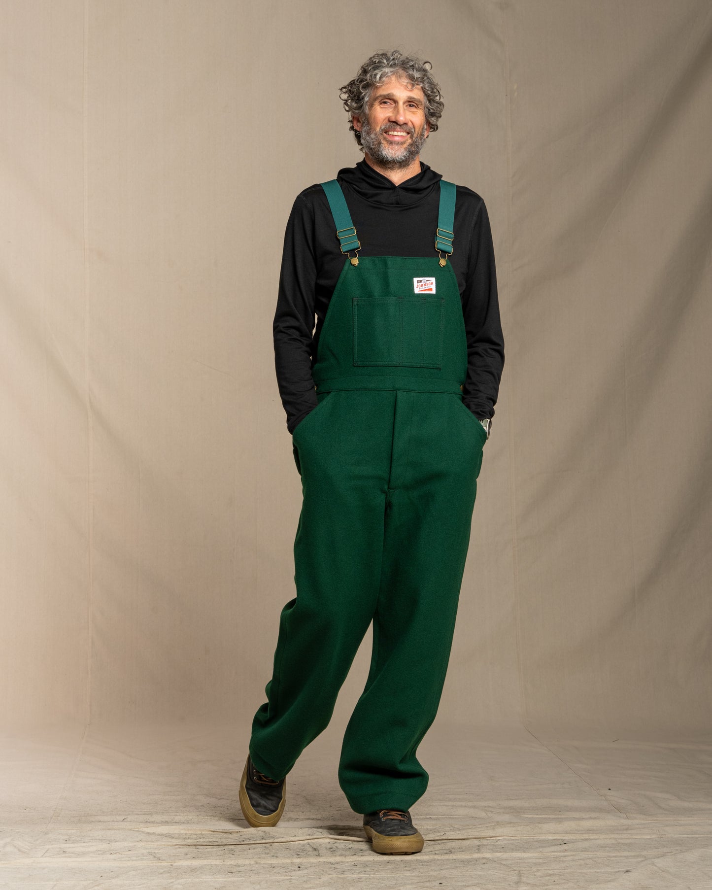 Man wearing green wool overalls and a black shirt against a plain background