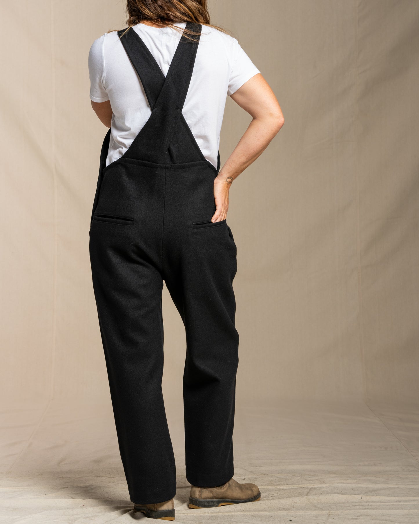 Person wearing black wool overalls and a white shirt against a beige background