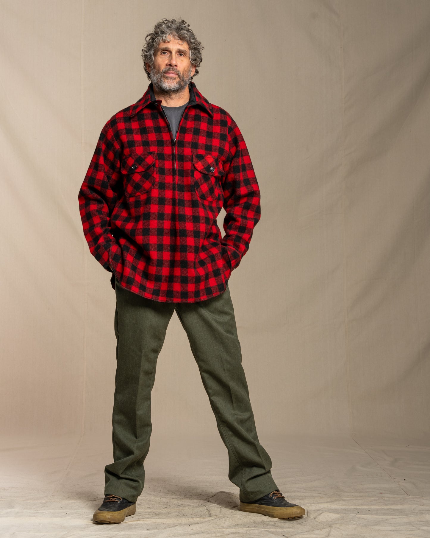 Man wearing a red and black checkered shirt and green pants against a beige background