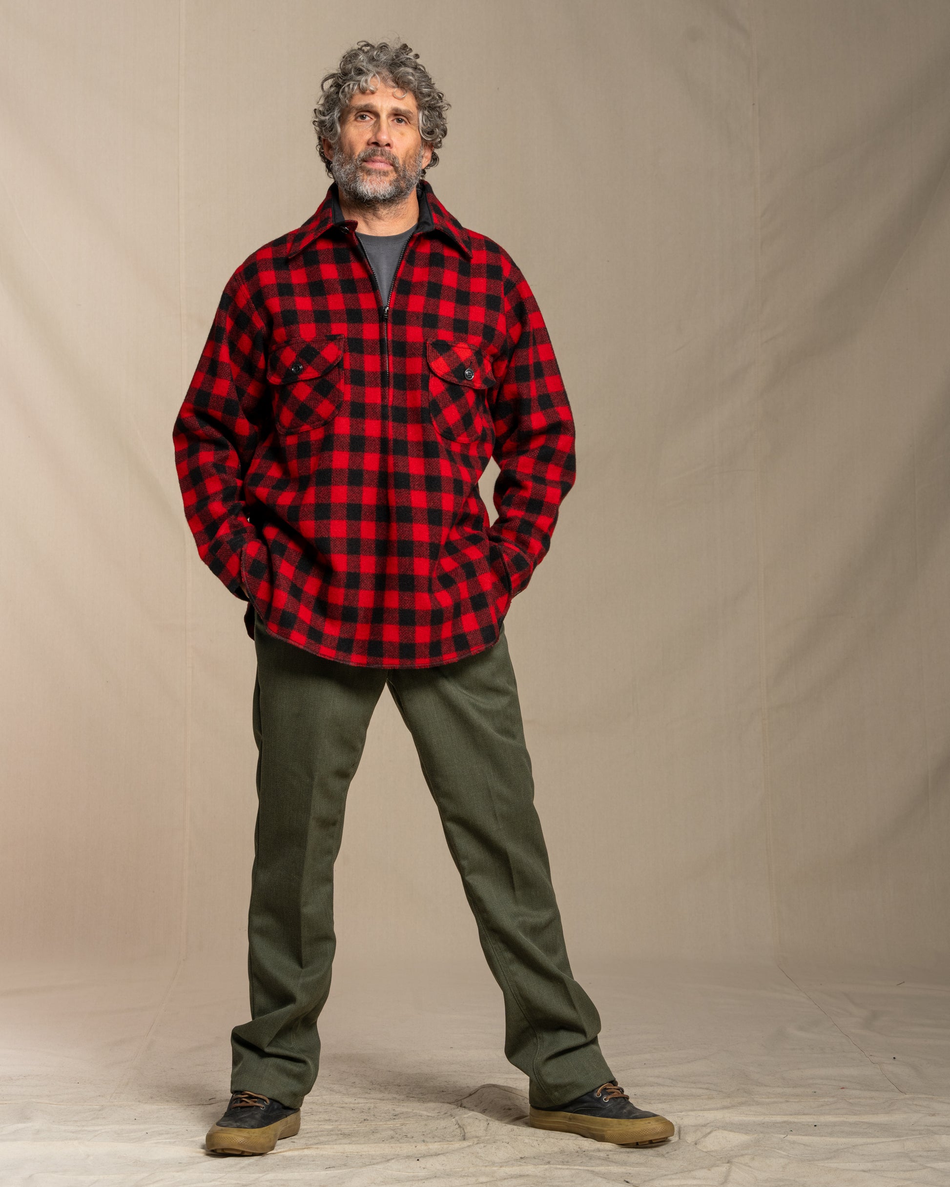 Man wearing a red and black checkered shirt and green pants against a beige background