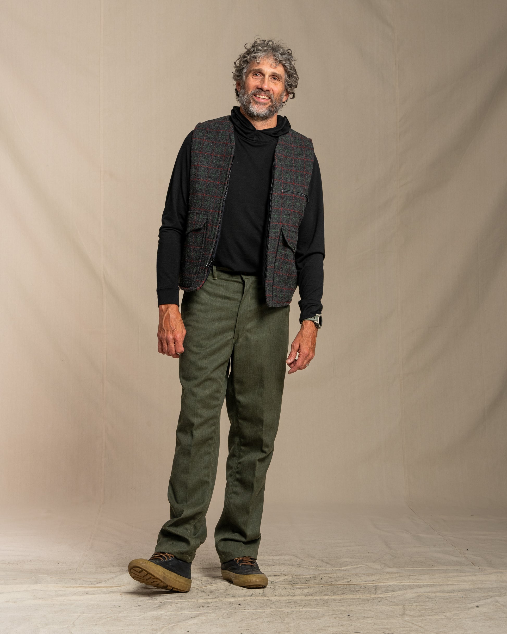 Man wearing a plaid wool vest, black shirt, and green pants standing against a beige background