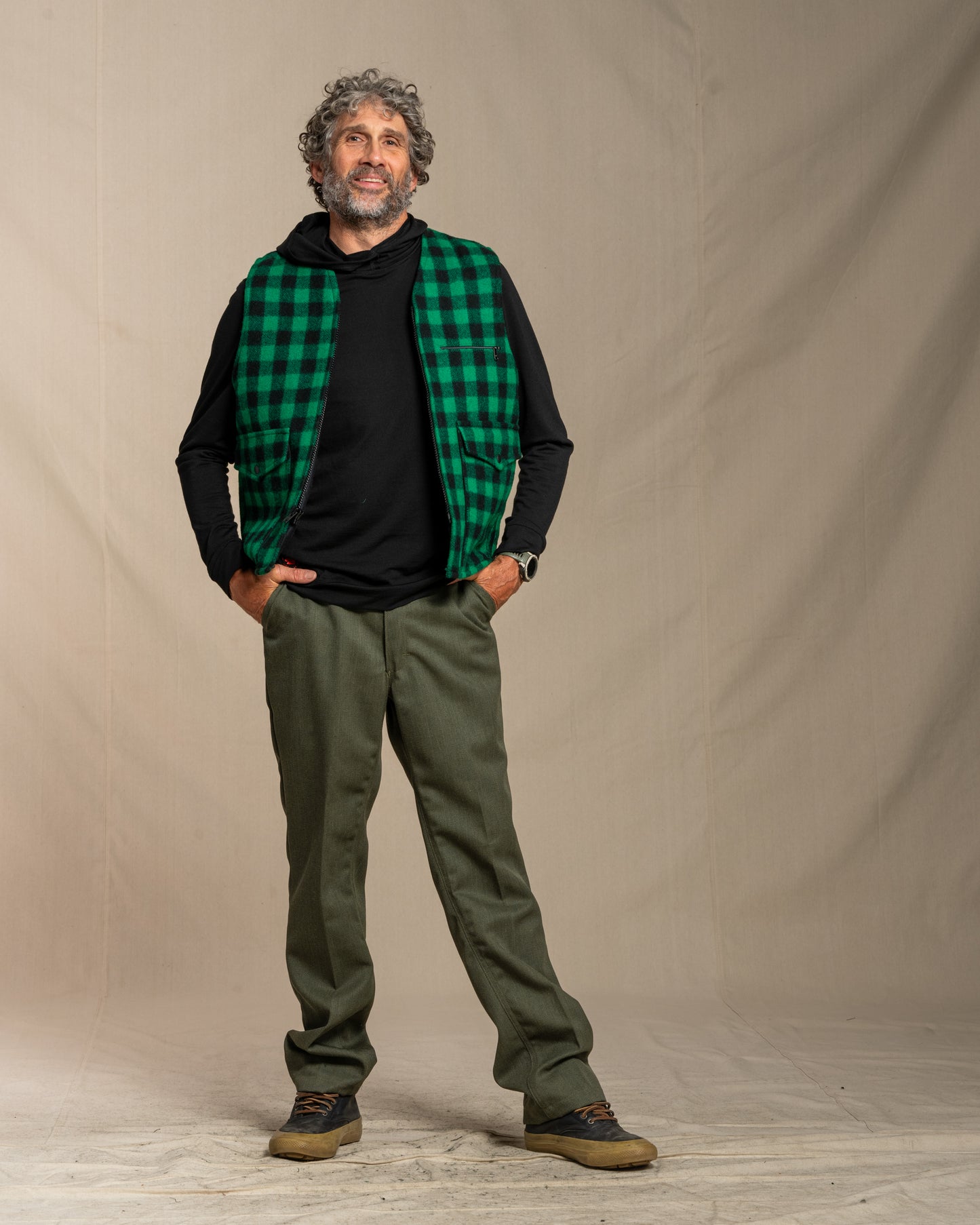 Man wearing a green plaid wool vest, black merino wool hoodie, and green wool pants against a beige background