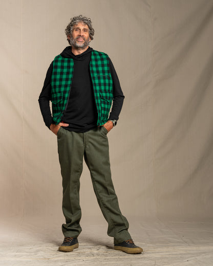 Man wearing a green plaid wool vest, black merino wool hoodie, and green wool pants against a beige background