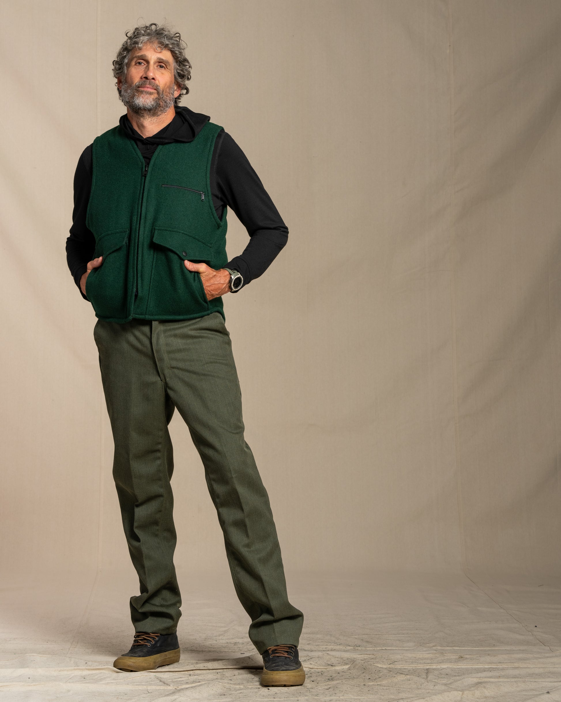 Man wearing a green wool vest over a black merino wool shirt with green wool pants against a plain background